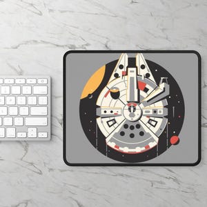 Könnte beinhalten: Eine graue Mausmatte mit schwarzem Rand, die eine stilisierte Illustration des Millennium Falcon Raumschiffs aus Star Wars zeigt. Das Raumschiff ist weiß mit grauen und roten Details. Der Hintergrund ist schwarz mit weißen Sternen und einem gelben und orangefarbenen Planeten.