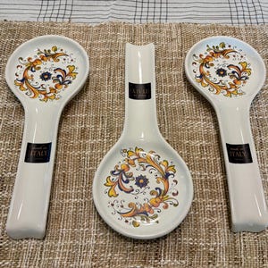 May include: Set of three white ceramic spoon rests with colorful floral designs in yellow, orange, and blue. The spoon rests are made in Italy and have a black label with white text.