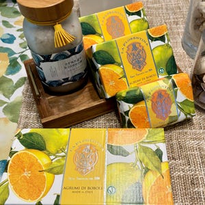 May include: A collection of bath products featuring an orange blossom theme. The image includes a jar with a wooden lid and tassel, and several boxes of soap with citrus fruit illustrations. The boxes are labeled "La Florentina" and "Agrumi di Boboli."