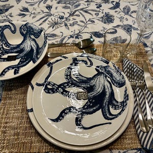 Peut inclure: Assiettes et un bol en céramique blanche avec un motif de poulpe bleu marine. Les assiettes sont empilées sur un dessous de plat tissé, avec des couverts, une serviette rayée et une nappe à motifs floraux. Les assiettes sont marquées "ITALY".