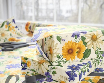 May include: Two gift boxes wrapped in floral patterned paper. The top box has a white background with yellow and purple flowers. The bottom box has a white background with yellow, purple, and green flowers. The boxes are sitting on a table covered in the same floral patterned paper.