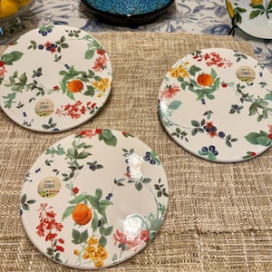 Puede incluir: Tres platos de cerámica blanca con diseños florales y frutales. Cada plato presenta una disposición diferente de flores rojas, amarillas y rosas, hojas verdes y frutas naranjas. Los platos tienen una etiqueta "Made in Italy".