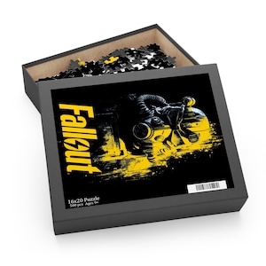 May include: A black puzzle box with the word "Fallout" in yellow. The box features a yellow and black graphic of a helmet and the text "16x20 Puzzle 500 pcs Ages 9+". The box is open, revealing puzzle pieces.