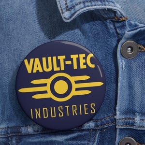 May include: A round, navy blue button with the words "VAULT-TEC INDUSTRIES" in yellow. The button features a yellow stylized atomic symbol. The button is pinned to a denim jacket.