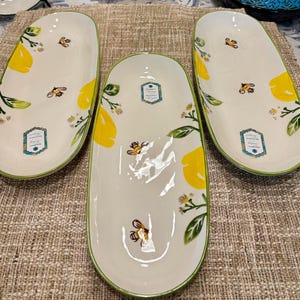 May include: Three white oval serving platters with a lemon and bee design. The platters have a green trim and feature yellow lemons, green leaves, and small bees. Each platter has a decorative label.