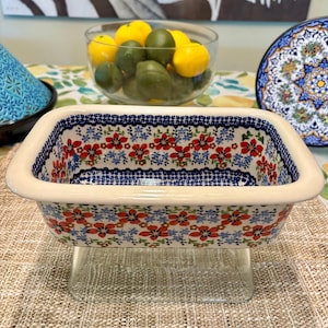 May include: Rectangular ceramic baking dish with a floral pattern in red, blue, and green on a cream background. The dish sits on a clear glass stand. The rim is trimmed with a blue and white design.