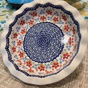 May include: A white ceramic pie dish with a scalloped edge. The dish features a blue and red floral pattern with a detailed blue center design. The rim is decorated with a blue border and the dish is sitting on a neutral-colored surface.