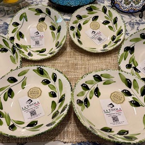 May include: A collection of handcrafted ceramic bowls with an olive branch design. The bowls are off-white with green and black olive branch patterns around the rims. Each bowl has a tag that reads "LA TAVOLA" and "ITALIE".
