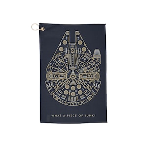 May include: A black golf towel featuring a gold outline of the Millennium Falcon spaceship. The text "What a piece of junk!" is printed below the spaceship.