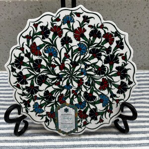 Puede incluir: Un posavasos de cerámica blanca con un diseño circular de flores rojas, azules y verdes. El posavasos tiene un borde festoneado y una etiqueta que dice "Handcrafted in Turkey. Fait à la main en Turquie."