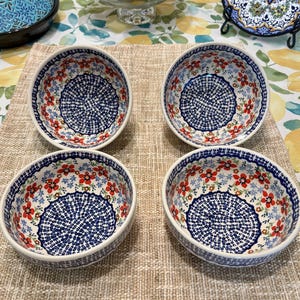 May include: Four ceramic bowls with a white base, adorned with a blue and red floral pattern. The bowls have a circular design in the center and a decorative border. The bowls are arranged on a tan surface.