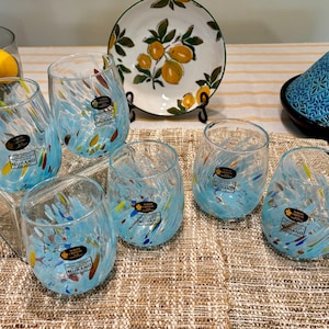 May include: Six blue glass tumblers with colourful flecks and a white label that reads "White Crystal Murano" on a woven placemat.