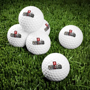 May include: White golf balls with a graphic design featuring skulls, a red flag, and the text "THE NEW BATCH". The golf balls are arranged on a bed of green grass.