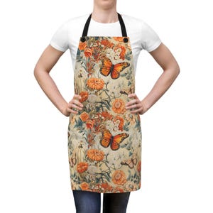Könnte beinhalten: Eine beige Schürze mit einem Blumenmuster mit orangefarbenen Schmetterlingen und Blumen. Die Schürze hat verstellbare Träger und eine Tasche.
