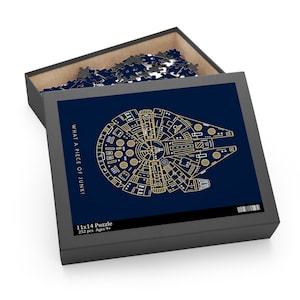 Puede incluir: Un rompecabezas de 252 piezas con un fondo azul y un contorno dorado del Halcón Milenario. El texto "What a piece of junk!" está impreso en la caja. La caja también indica "11x14 Puzzle" y "Ages 9+".