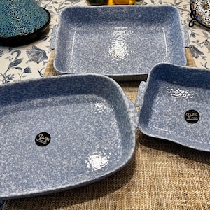 Puede incluir: Tres fuentes de horno de cerámica azul claro con un patrón moteado. Las fuentes son rectangulares con esquinas redondeadas y asas. Cada fuente tiene una pegatina en blanco y negro. Las fuentes están sobre un mantel estampado.