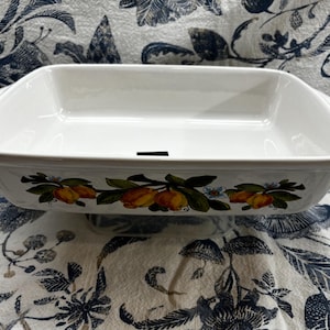 Puede incluir: Plato para hornear de cerámica blanca con un diseño floral con frutas amarillas y naranjas. El plato tiene un borde ligeramente elevado y dos asas.