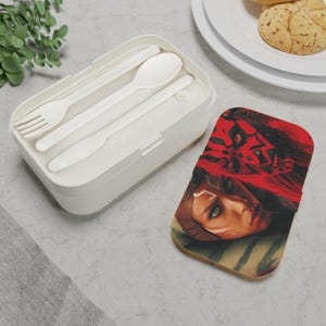 May include: A white rectangular lunch box with a fork, spoon, and knife inside. A coaster with a red and black graphic of a character's face is next to the lunch box. The lunch box is on a light gray surface.