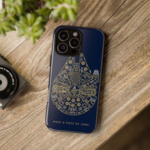 Op de afbeelding: Een donkerblauw smartphonehoesje met een goudkleurige lijntekening van een ruimteschip. Het ontwerp bevat ingewikkelde mechanische details en de tekst WHAT A PIECE OF JUNK! in goud gedrukt aan de onderkant van het telefoonhoesje.