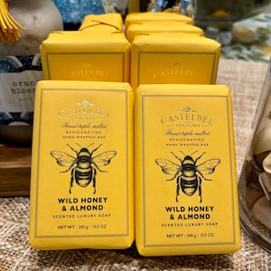 May include: Yellow Castelbel soap bars with a bee illustration and the words "Wild Honey & Almond." Each bar is hand-wrapped and weighs 10.5 oz. The soap is triple-milled and scented. The packaging is yellow.