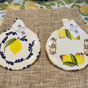 May include: Two round, hand-painted ceramic serving platters with a handle. One features a lemon design with blue accents, the other has lemons and brown decorative swirls. Both have tags that read "LA TAVOLA Handpainted Ceramics Made in Italy."