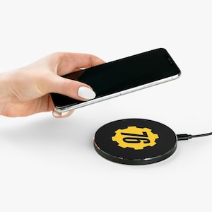 May include: A black wireless phone charger with a yellow gear and the number 76 printed on it. The charger is connected to a black cable. A black smartphone is held above the charger.