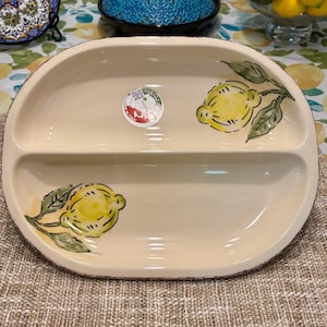 May include: Cream-colored, oval-shaped ceramic serving dish with two compartments. The dish is decorated with hand-painted yellow lemons and green leaves. A small sticker with text is visible on the dish. The rim has a brown, distressed edge.