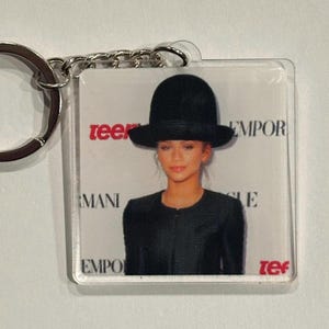 May include: A clear acrylic keychain featuring a photograph of a person wearing a black hat and black clothing. The keychain has a silver metal ring and chain. The word "teen" is visible in red.