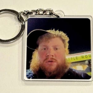 May include: Square acrylic keychain with a silver metal ring. The keychain features a photograph of a person with a beard and a hat. The background is dark, with the text "WAFFLE HOU" visible.