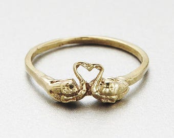 Könnte beinhalten: Ein goldfarbener Metallring mit zwei Schwänen, die einander zugewandt sind. Die Hälse der Schwäne krümmen sich nach oben und bilden einen herzförmigen Raum zwischen ihnen. Der Ring ist glatt und rund und mit der Basis der Schwäne verbunden.