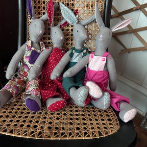 May include: Four gray bunny dolls with long ears, each wearing different outfits. One wears a floral print dress, another a red polka-dot dress, a third a green overall, and the last a pink overall. They are seated on a woven chair.