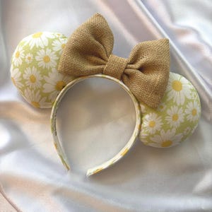 May include: A headband with two round ears and a large bow. The ears are covered in a light green fabric with a white daisy pattern. The bow is made of a tan burlap material. The headband is white with a daisy pattern.