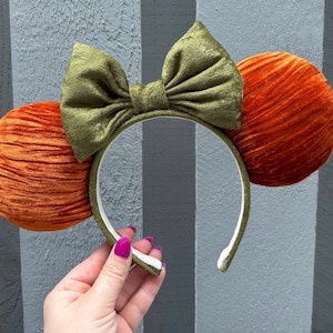 May include: A headband with two orange, round ears and a green bow. The ears and bow are made of a textured fabric. The headband is white on the inside and green on the outside.