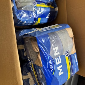 以下が含まれることがあります： TENA MENの保護用下着が複数入った段ボール箱。 パッケージは青と白で、「MEN」の文字が大きく表示されています。 下着はグレーとブルーのようです。