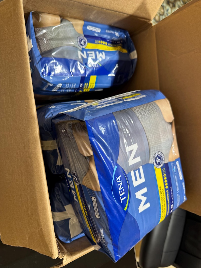 以下が含まれることがあります： TENA MENの保護用下着が複数入った段ボール箱。 パッケージは青と白で、「MEN」の文字が大きく表示されています。 下着はグレーとブルーのようです。