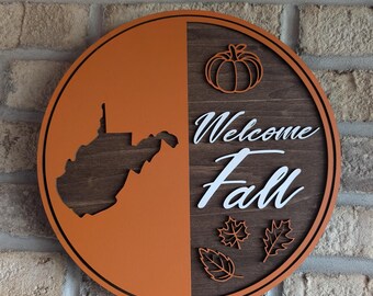 May include: A round wooden sign with an orange background and a brown wood grain background. The sign features a silhouette of the state of West Virginia on the orange side and the words "Welcome Fall" with a pumpkin and leaves on the wood grain side.