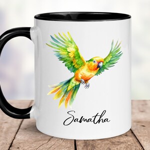 May include: White ceramic mug with a black rim and handle. The mug features a watercolour illustration of a green and yellow parrot in flight. The name "Samantha" is written in cursive at the bottom of the mug.