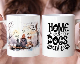 May include: White ceramic mug with a watercolor illustration of a woman and two dogs in a snowy forest. The text "Home is where the dogs are" is printed on the other side of the mug.
