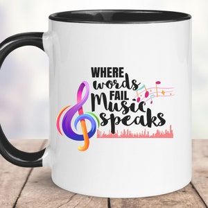 Peut inclure: Une tasse à café en céramique blanche avec une poignée noire et un bord noir. La tasse présente une clé de sol colorée et des notes de musique avec le texte "Where words fail music speaks".