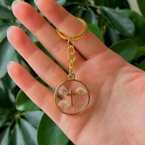 Puede incluir: Un llavero dorado con un colgante circular. El colgante contiene una peque&ntilde;a cruz dorada y delicadas flores blancas encapsuladas en resina transparente. El llavero se sostiene frente a una exuberante planta verde.