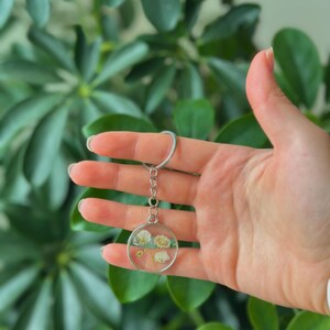 Puede incluir: Un llavero redondo transparente con flores blancas secas encapsuladas en resina. El llavero tiene un marco y una cadena de metal plateado, y un llavero plateado. El fondo es una exuberante planta verde.