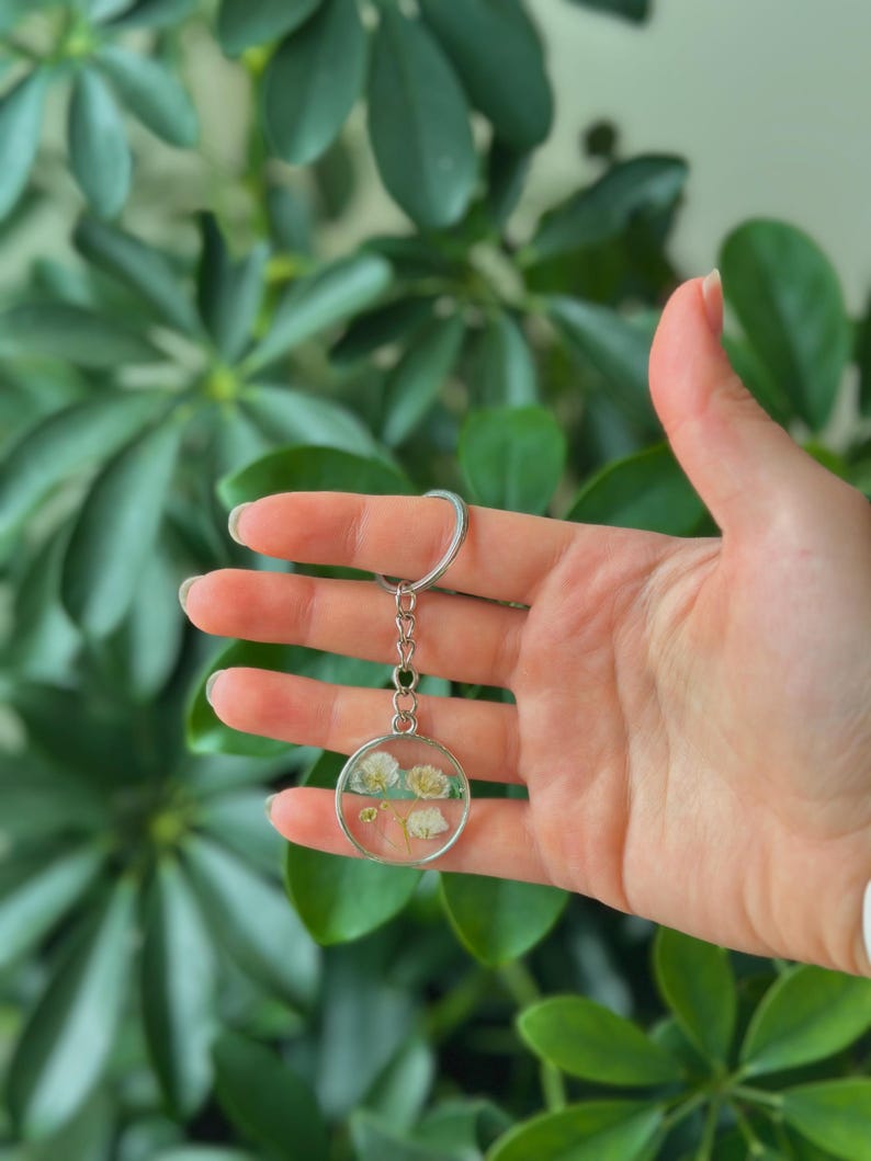 Puede incluir: Un llavero redondo transparente con flores blancas secas encapsuladas en resina. El llavero tiene un marco y una cadena de metal plateado, y un llavero plateado. El fondo es una exuberante planta verde.