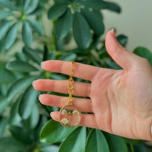 Puede incluir: Un llavero dorado con un colgante redondo. Dentro del colgante hay peque&ntilde;as flores blancas y una cruz dorada. El llavero se sostiene frente a un fondo de hojas verdes.