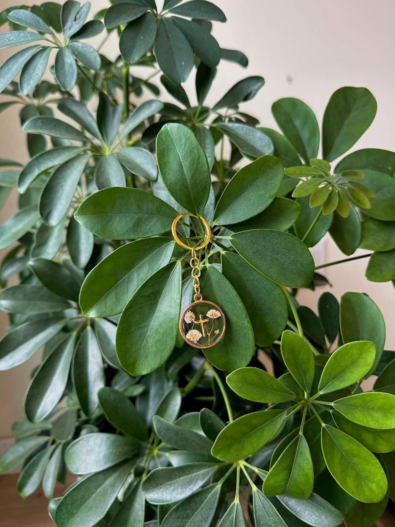 Puede incluir: Un llavero dorado con un colgante redondo con flores secas y un dise&ntilde;o de cruz. El llavero cuelga sobre un fondo de hojas verdes exuberantes.