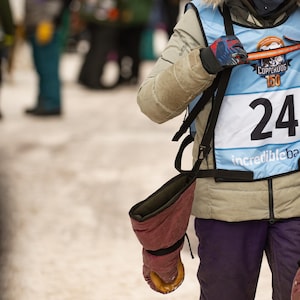 Puede incluir: Una persona joven que lleva un chaleco azul y blanco con el número 24. El chaleco tiene el texto "Copperdog 150" e "Incredible Bay". La persona lleva una chaqueta marrón con capucha y ribete de piel y sostiene un palo naranja largo.