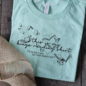 May include: A light teal t-shirt with the text "this is my binge reading shirt" and the quote "if my book is open, your mouth should be shut." The shirt features book and butterfly graphics.
