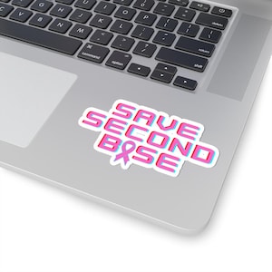 Peut inclure: Un autocollant blanc avec le texte "SAVE SECOND BASE" en rose et bleu, avec un ruban rose. L'autocollant est sur un ordinateur portable argenté.