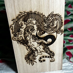 May include: Wooden plaque featuring a detailed pyrography design of a dragon and tiger intertwined around a yin-yang symbol. The artwork is burned into the light-colored wood, creating a striking contrast.