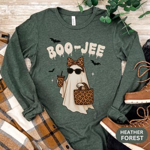 May include: A dark green long-sleeve t-shirt featuring a graphic of a ghost wearing a leopard print bow and sunglasses, holding a leopard print cup and tote bag. The text BOO-JEE is printed above the ghost in a white, dripping font style.