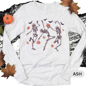 May include: A light gray long-sleeve shirt featuring a graphic print of three dancing skeletons. The skeletons are surrounded by small black bats and orange autumn leaves. One skeleton holds a small orange pumpkin, and another carries a bucket.